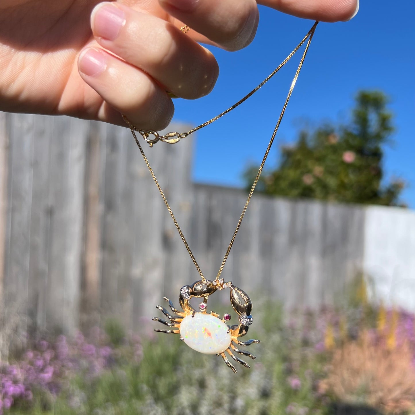 A gold opal crab necklace being dangled outdoors in natural sunlight, revealing a bright red opal flash.