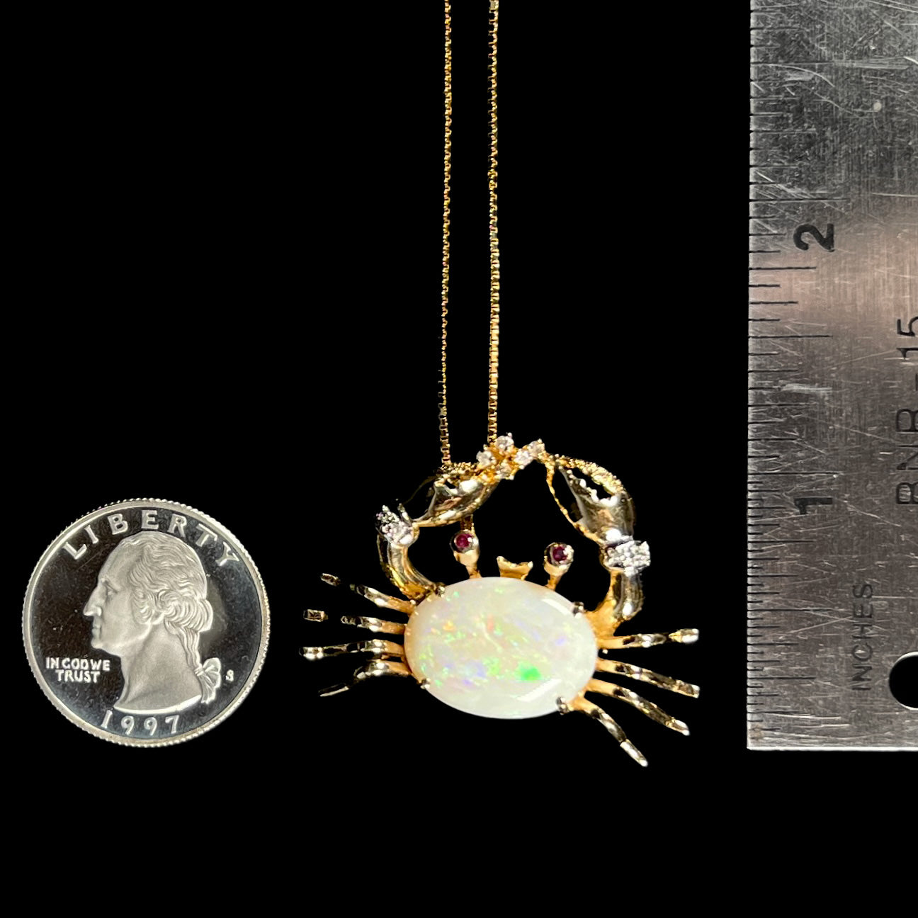 Gold opal crab necklace beside a quarter and ruler for scale, measuring approximately 1.25 inches long, with green opal flashes.