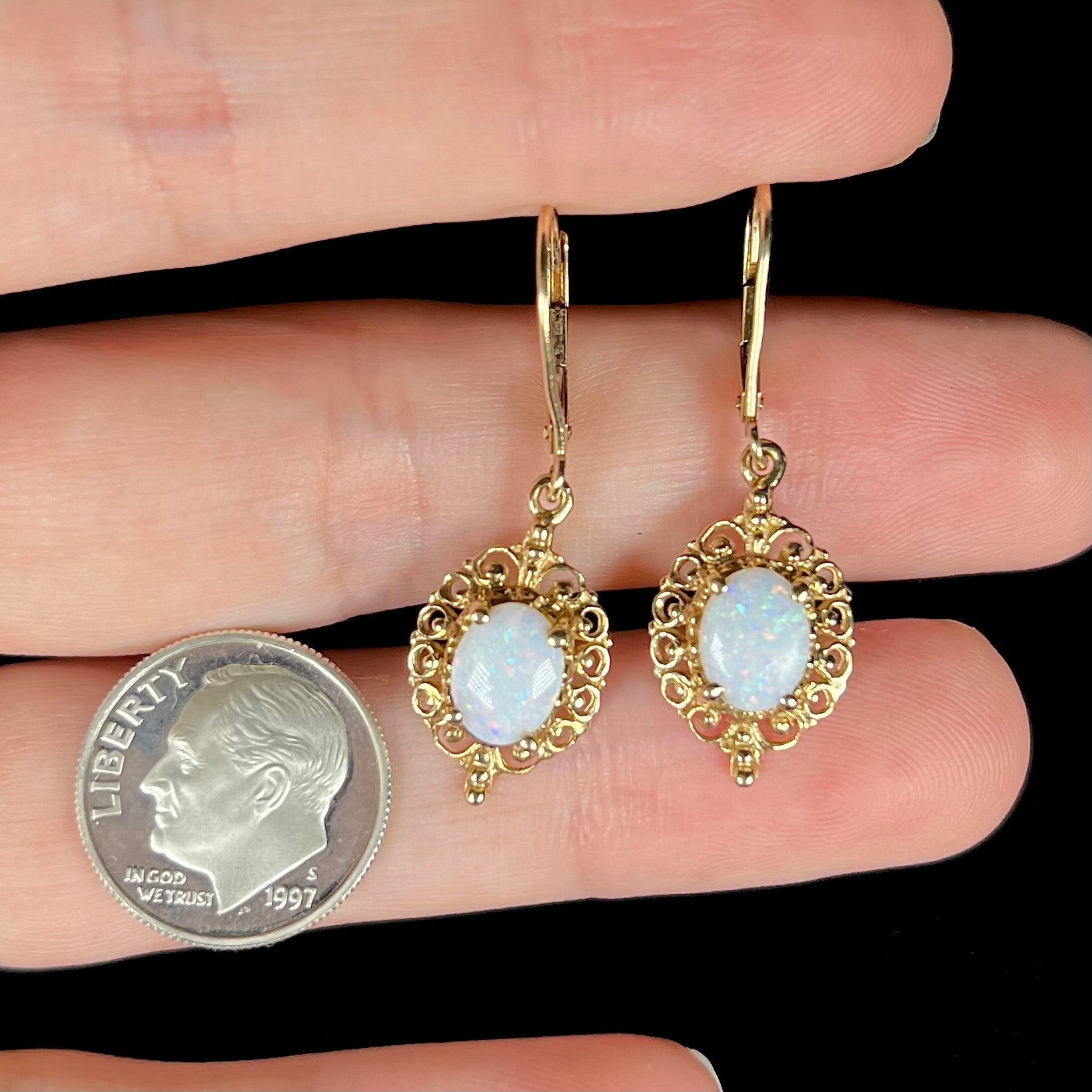 A pair of vintage yellow gold Australian light opal dangle earrings hown close-up in a hand next to a dime coin.