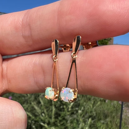 Pair of gold opal drop earrings held between two fingers at angle in natural light against green utdoor background.
