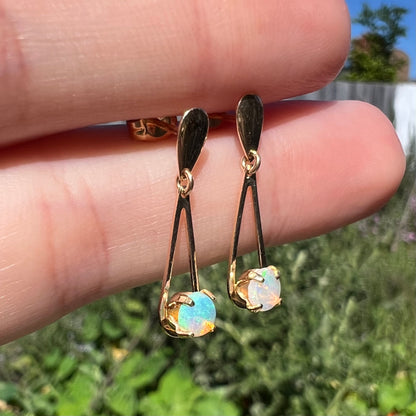 Close-up angled view of yellow gold opal drop earrings held between two fingers in sunlight against blurred leafy background.