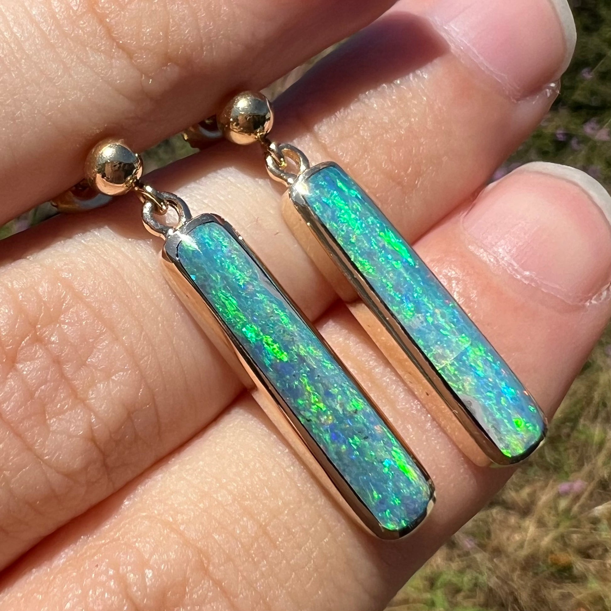 Yellow gold opal drop earrings with green color play, shown at an angle in natural sunlight.