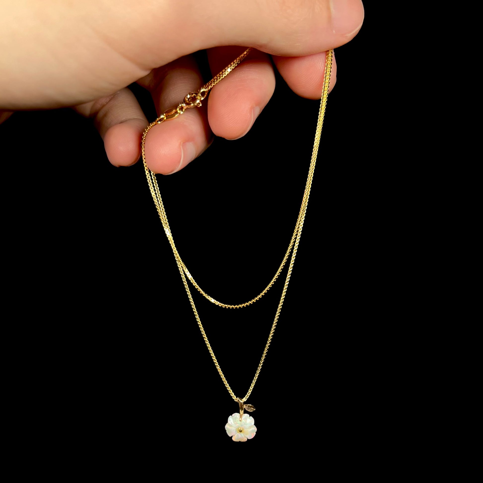 A gold opal flower necklace shown dangled by a hand from its chain against a black background.