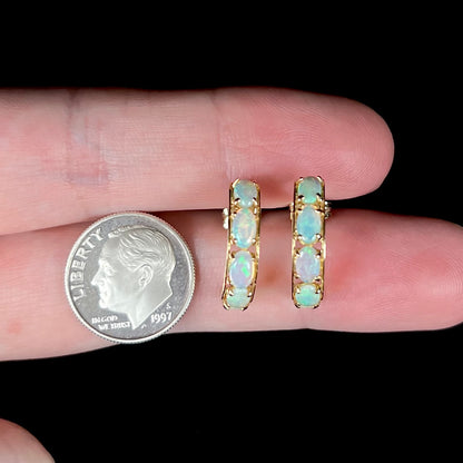 A pair of solid gold opal hoop earrings held between two fingers next to a US dime coin.