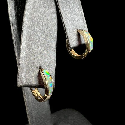 A pair of yellow gold lab-created opal inlay huggie hoop earrings shown from an angle against a black background on an earring display.