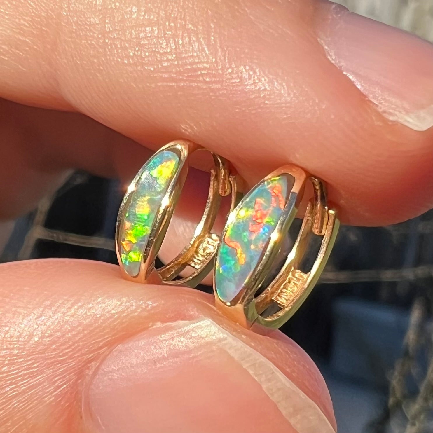 A pair of yellow gold lab-created opal inlay huggie hoop earrings shown in the sunlight from a close-up angle between two fingers.