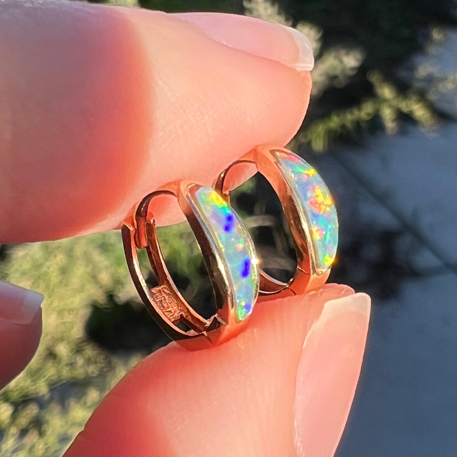 Close-up angled view of a pair of yellow gold lab-created opal inlay huggie hoop earrings held between two fingers in sunlight.