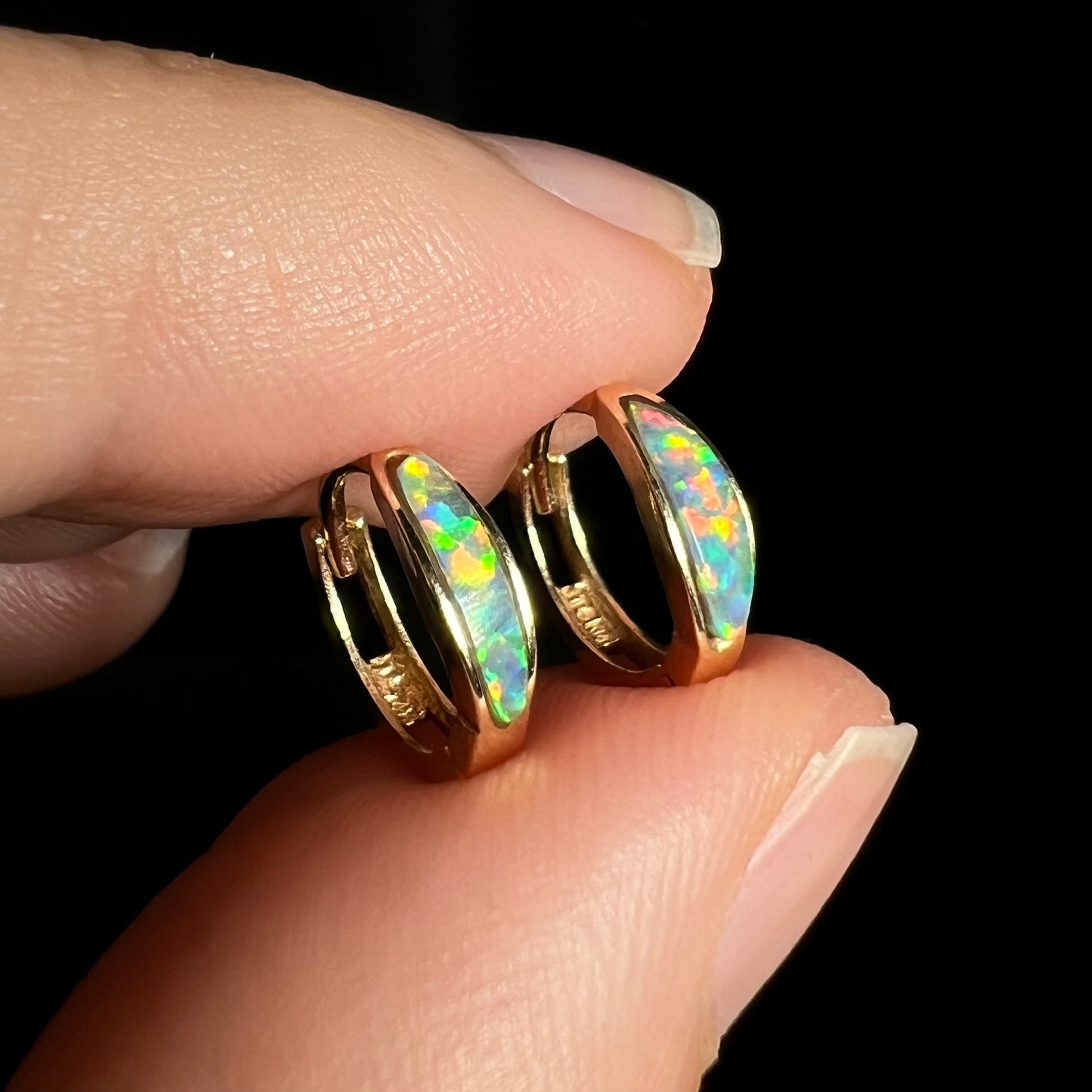 Close-up angled view of a pair of yellow gold lab-created opal inlay huggie hoop earrings shown between two fingers against a black background.