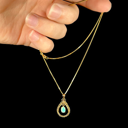 An 18k gold opal necklace being dangled by the chain in a hand against a black background, showing a greenish hue in the opal.