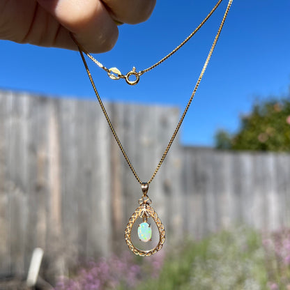 A gold opal filigree necklace being dangled outdoors in sunlight, with only subtle greenish colors visible.