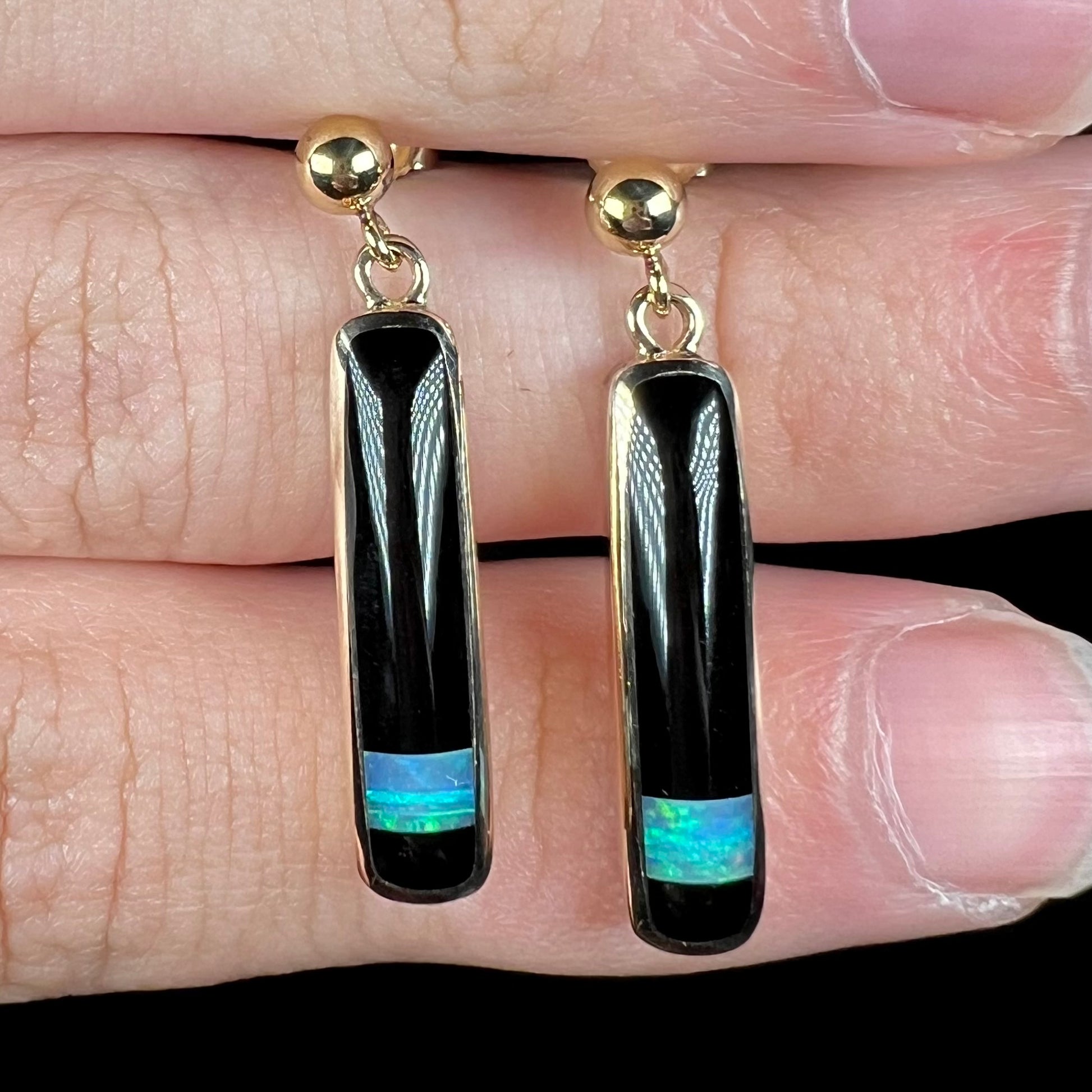 Close-up front view of a pair of gold opal onyx inlay earrings, held on top of rows of fingers.