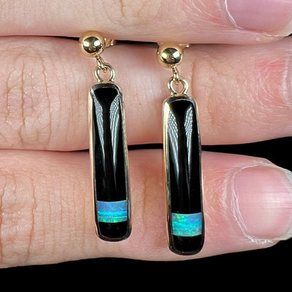 Close-up front view of a pair of gold opal onyx inlay earrings, held on top of rows of fingers.