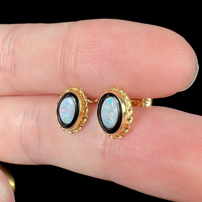 Yellow gold opal and onyx stud earrings with rope design bezels shown close-up from angle between two fingers under LED studio light.