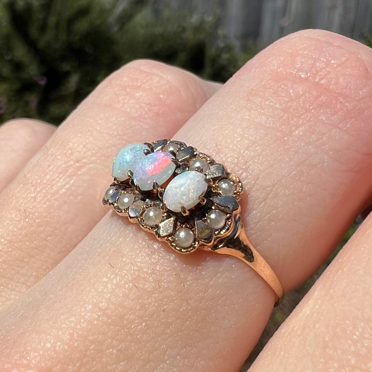 Angled close-up of 9k gold opal and seed pearl ring, worn on finger in sunlight
