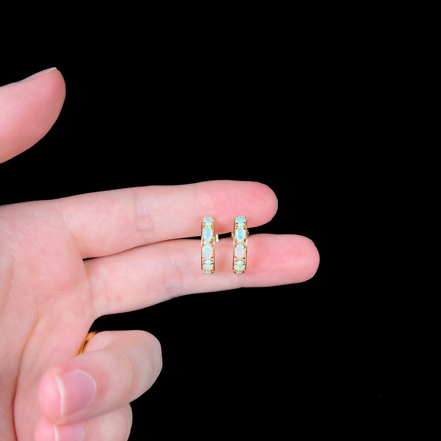 A pair of yellow gold opal semi-hoop earrings held between two fingers of a hand against a black background.