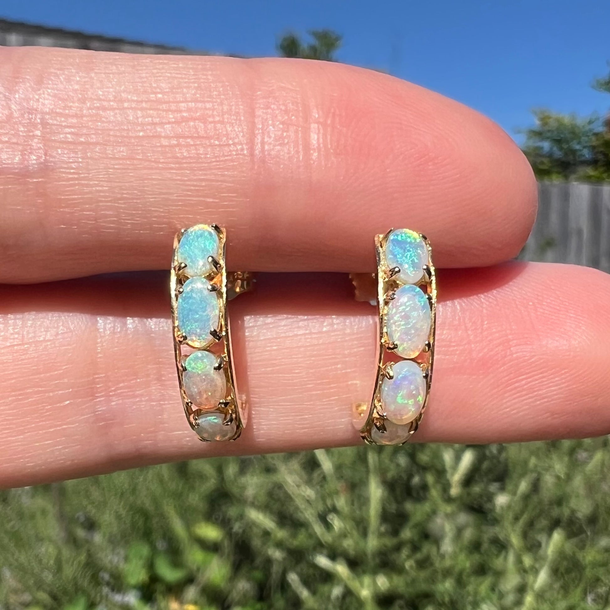 Close-up of a pair of gold opal semi-hoop earrings held between two fingers in natural light against a blurred outdoor background.