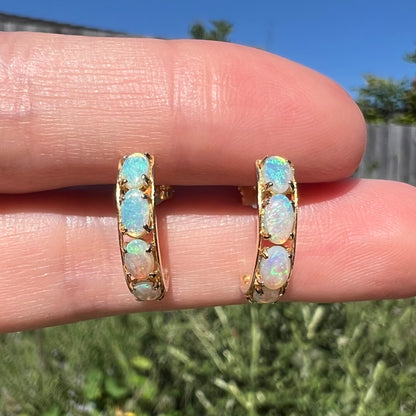 Close-up of a pair of gold opal semi-hoop earrings held between two fingers in natural light against a blurred outdoor background.