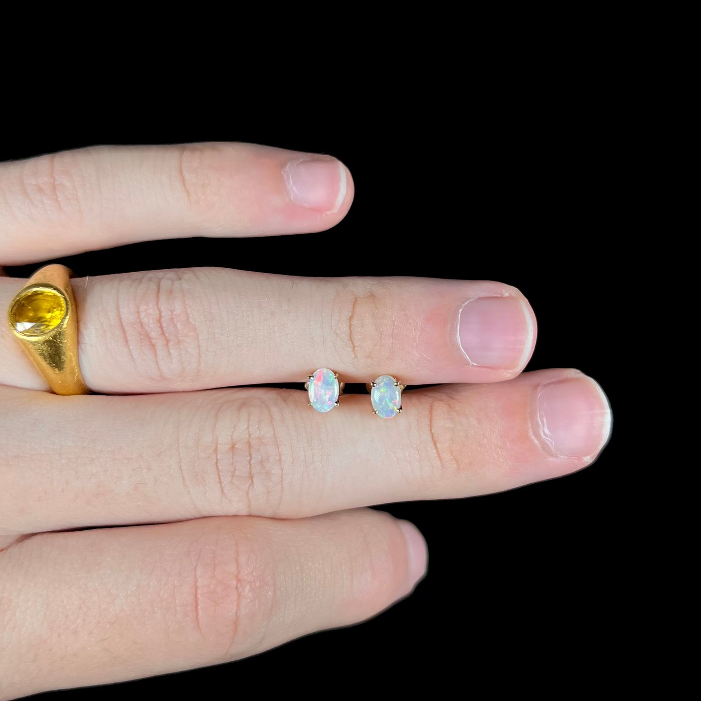 Gold opal stud earrings held between fingers, worn with a yellow sapphire gold ring.