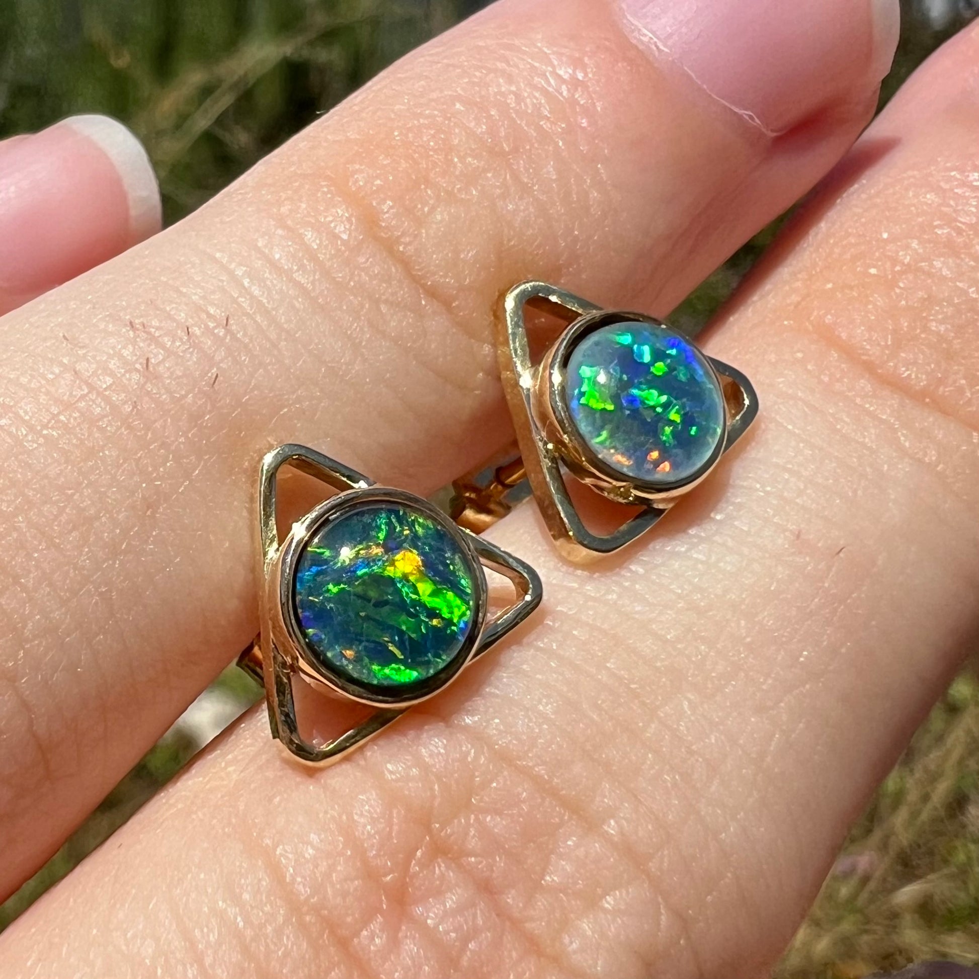 Angled sunlit view of a pair of gold opal triplet earrings with an open triangle design, shown on a hand.