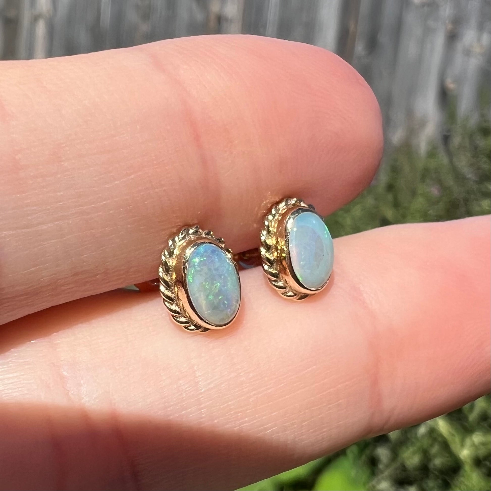 Gold opal earrings with rope bezel design shown from side close-up between two fingers in sunlight.