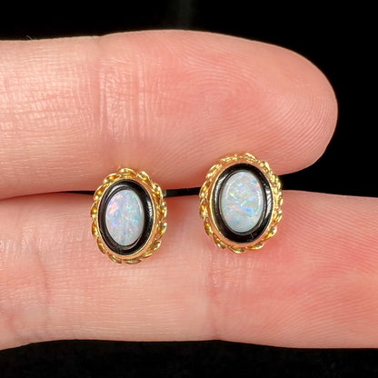 Close-up of gold rope design opal in onyx stud earrings held between two fingers under LED studio lights.