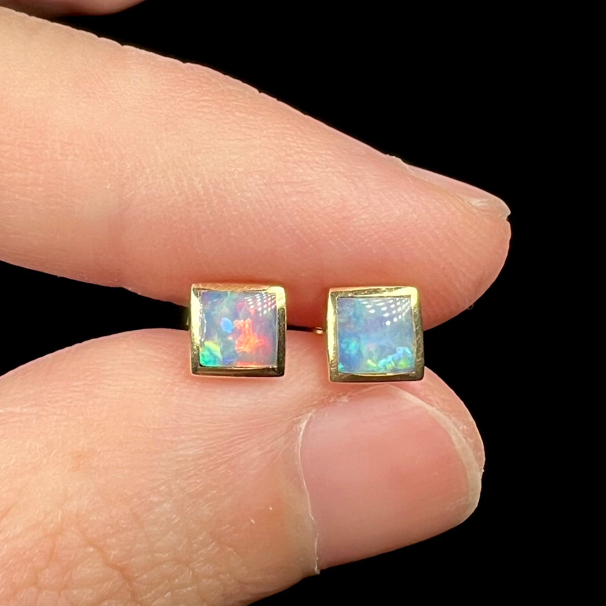 Close-up of 14k gold stud earrings with square shaped opals, held in fingers