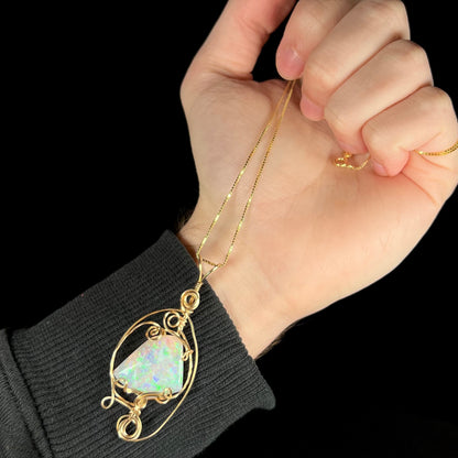 Hand dangling a gold-tone wire wrapped necklace set with a natural Lambina opal from its box-style chain.  The pendant rests against the person's wrist.