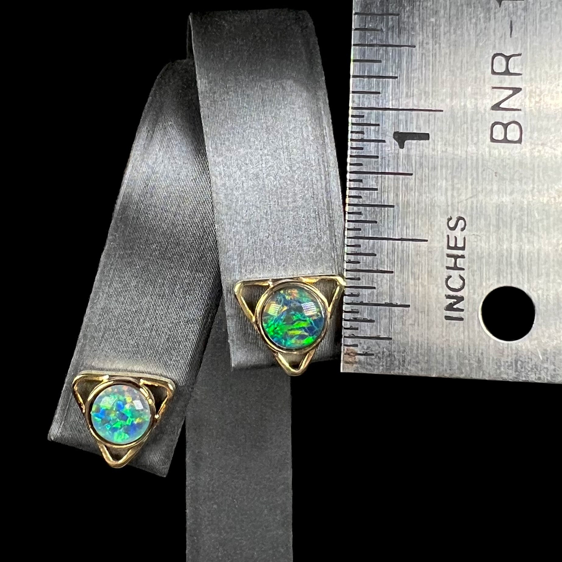 A pair of gold triangle earrings with round black opal triplets on a display next to a ruler, measuring 0.33 inches.