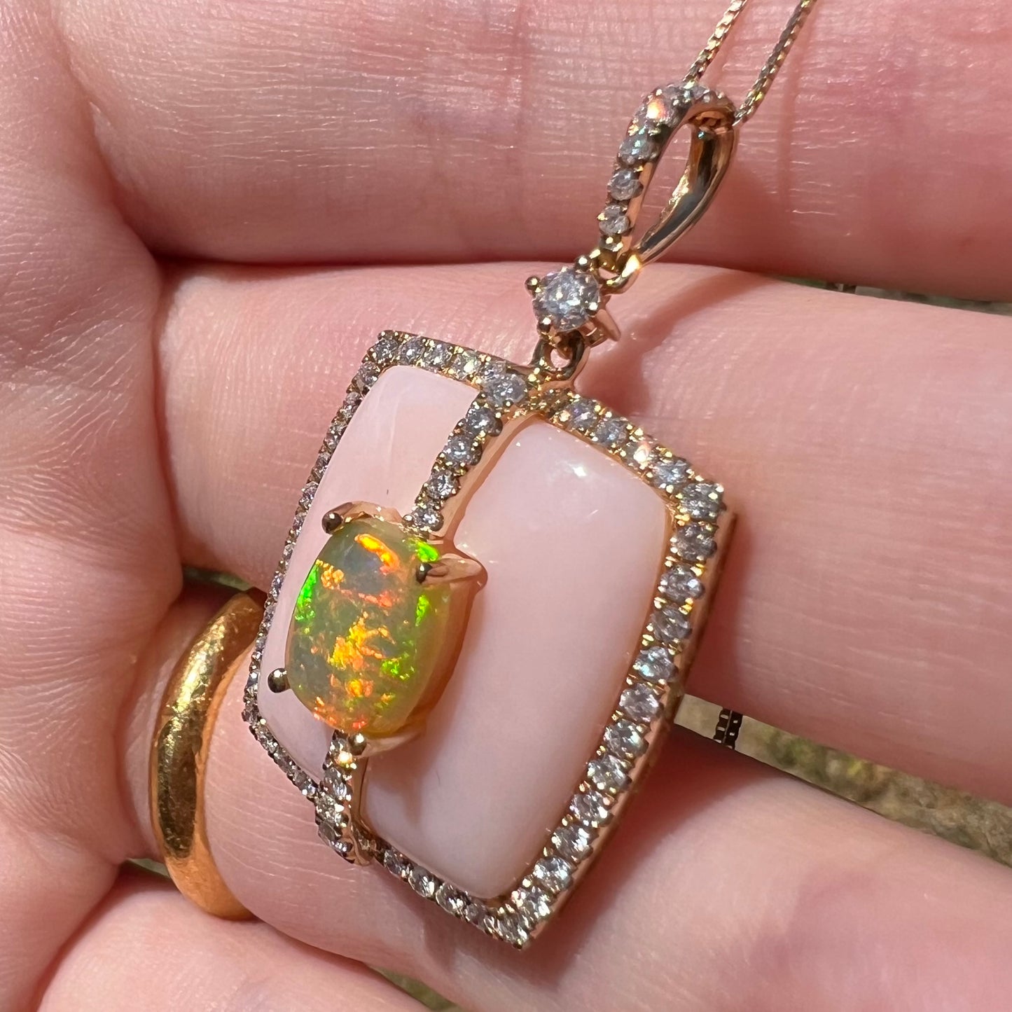 A women's gold Ethiopian fire opal and diamond pendant with a pink Peruvian opal backdrop shown close-up in sunlight, held in a hand.