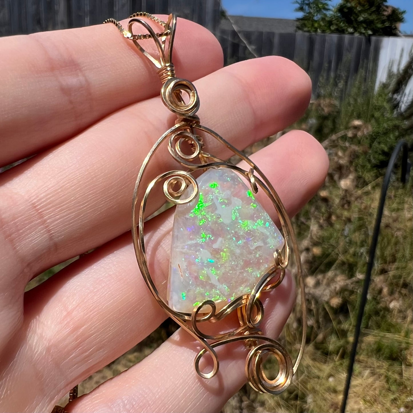 Angled view of a wire wrapped Australian crystal opal pendant held in sunlight, green flashes of color visible across the opal's surface.