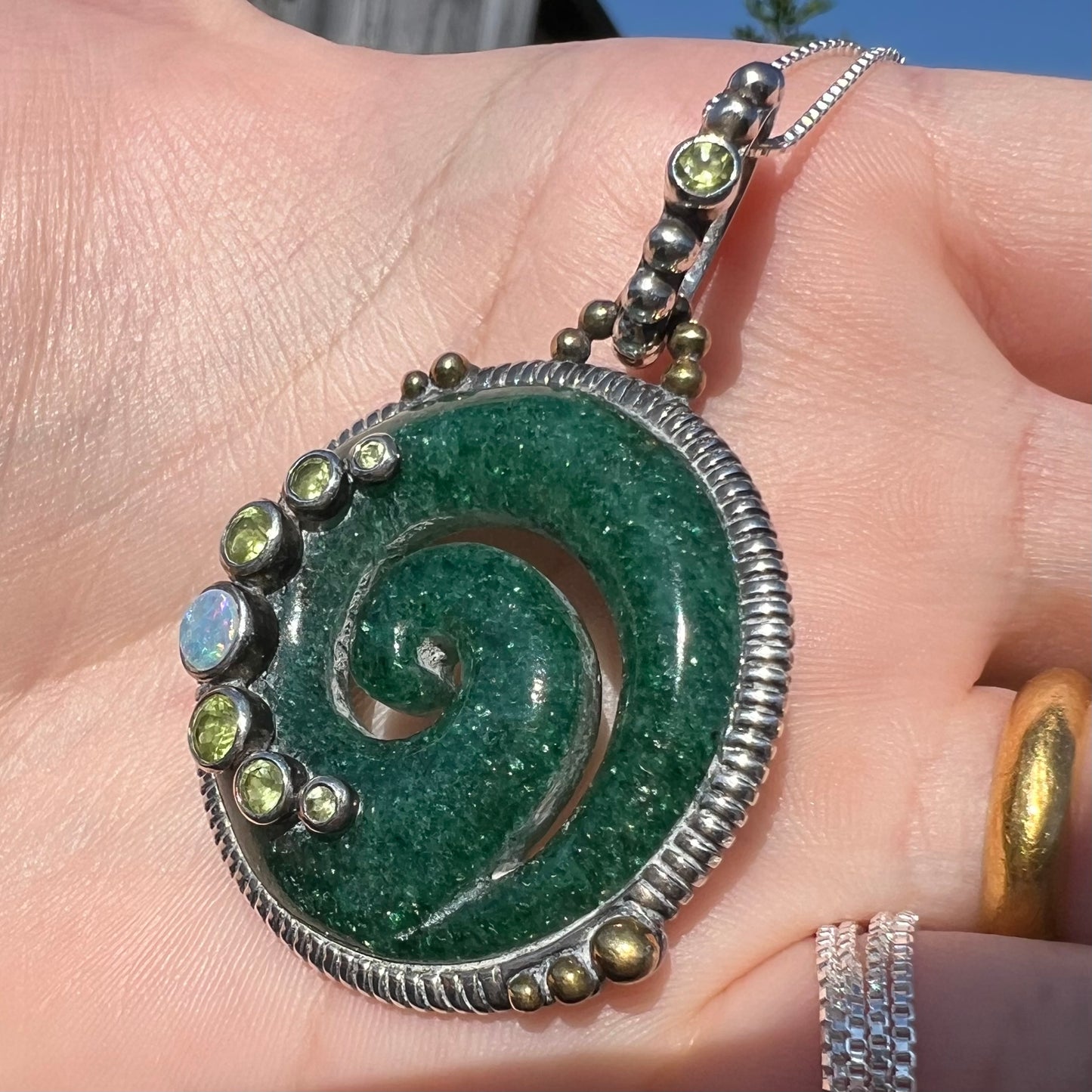 A men's aventurine koru necklace, accented with opal and peridot, shown in the sunlight from an angle.