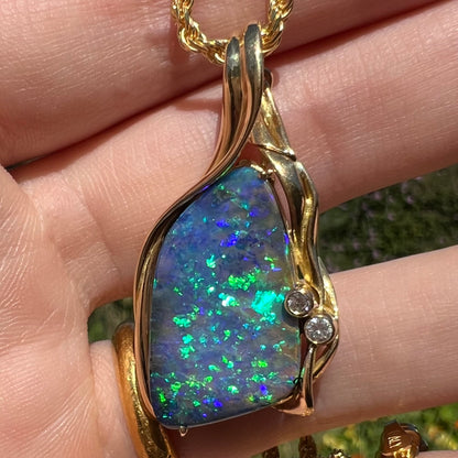 Close-up front view of green and blue Australian boulder opal pendant with diamond accents held in hand under natural sunlight.