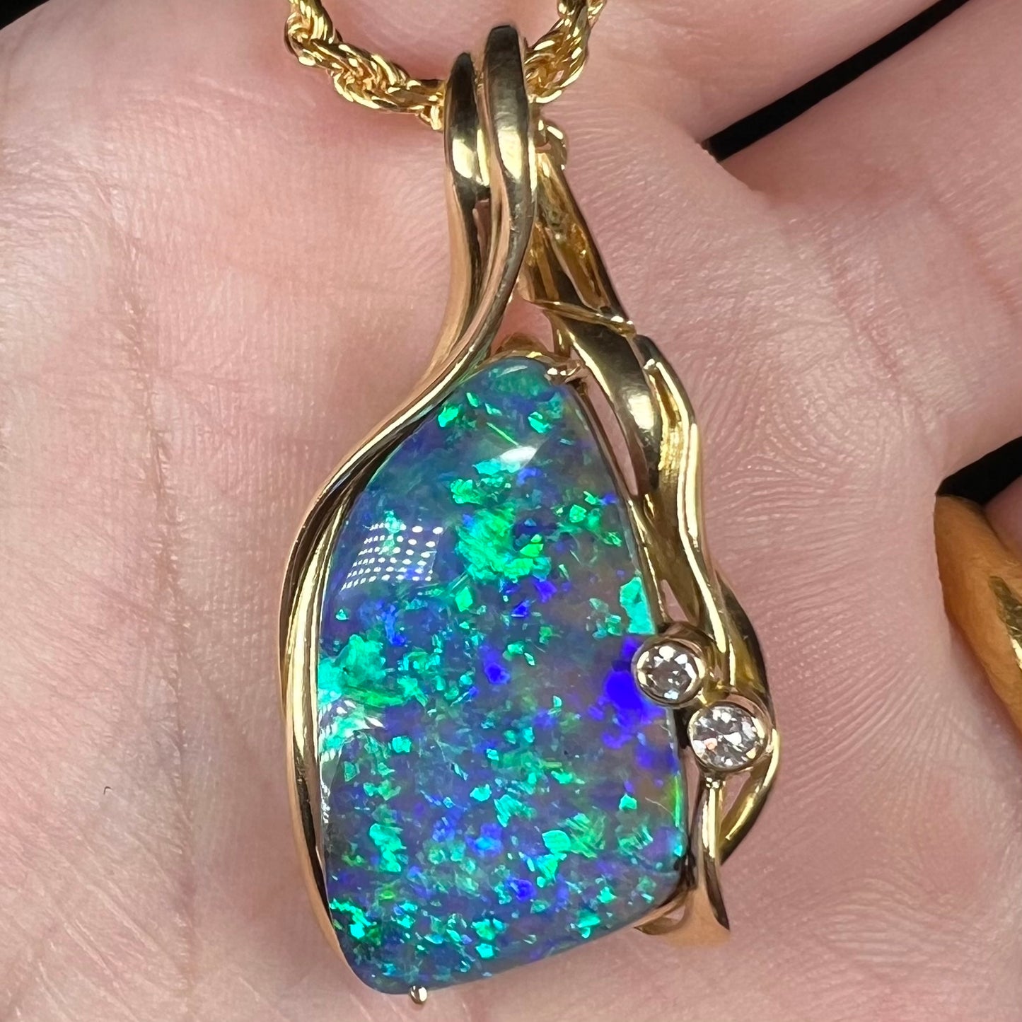 Close-up macro view of Australian boulder opal and diamond pendant with green-blue fire, held in hand.