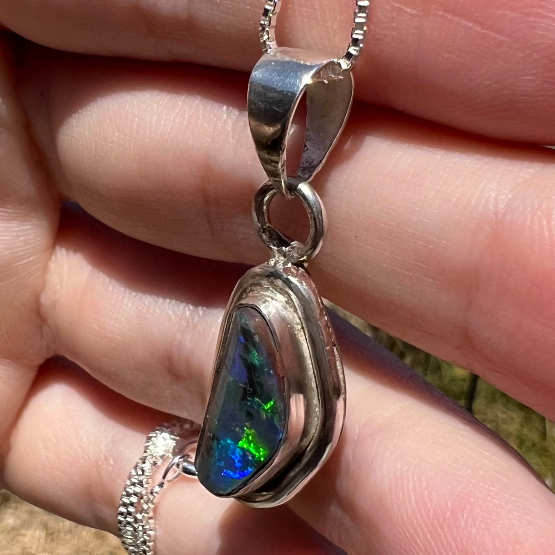 Green and blue flashes visible from a sterling silver black opal pendant, shown close-up from an angle in a hand under sunlight.