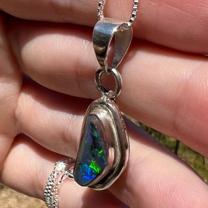 Green and blue flashes visible from a sterling silver black opal pendant, shown close-up from an angle in a hand under sunlight.