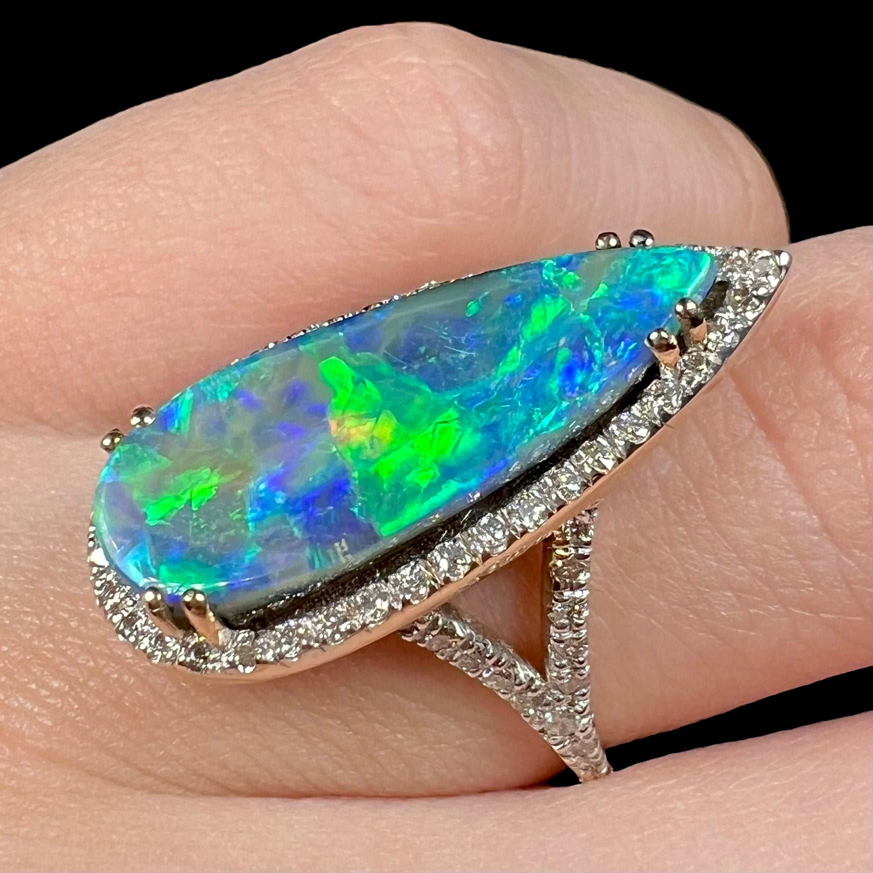Angled close-up of a pear-shaped boulder opal and diamond halo ring with green and blue color play, worn on a finger.