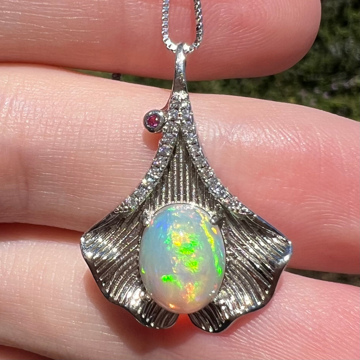 Close-up of iridescent green Ethiopian opal pendant with sterling silver ginko leaf design, held in hand under sunlight