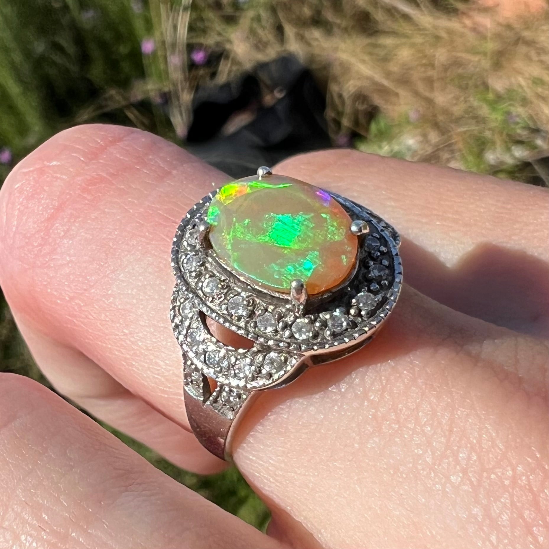 Angled close-up of a bluish green Ethiopian opal ring with a halo of CZs, worn on a finger in the sunlight.