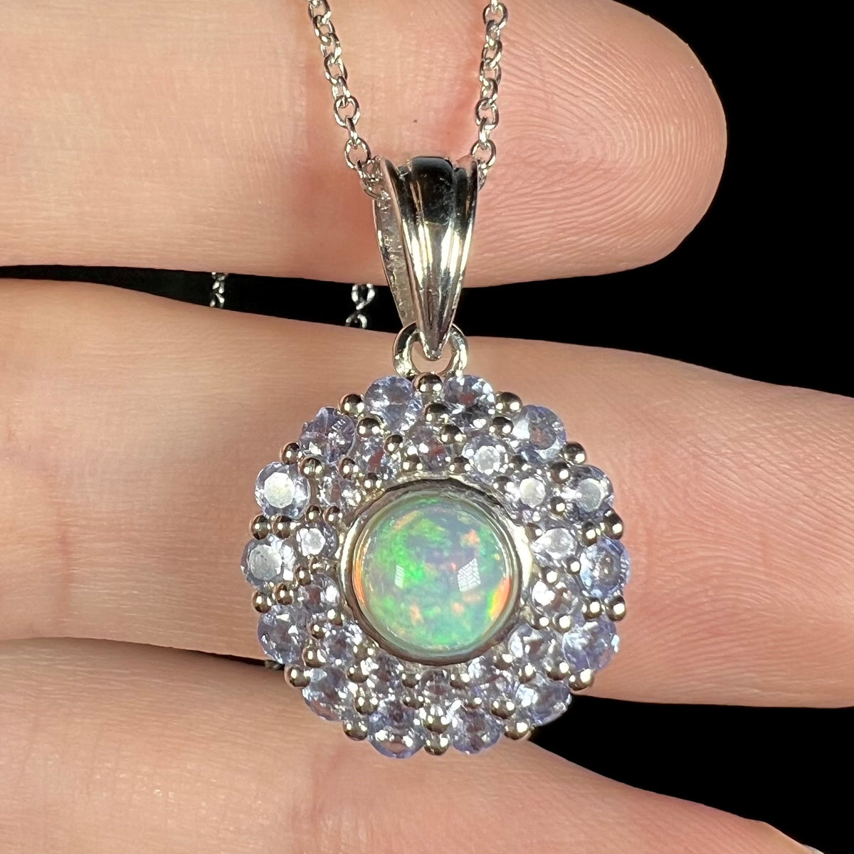 Macro close-up of a green Ethiopian fire opal and tanzanite halo pendant in sterling silver, shown resting on fingers.