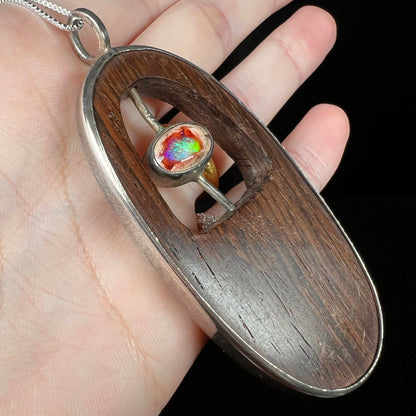 Angled close-up of a Mexican cantera fire opal and carved brown wood men's pendant in sterling silver, held in a hand.  The opal has a bright green flash of color.