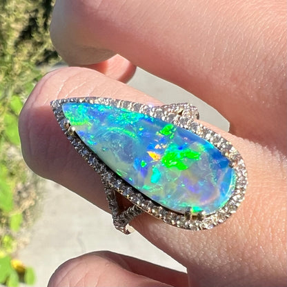 Sunlit close-up of a pear-shaped boulder opal and diamond halo ring with a bright green flash of color play, worn on a finger.