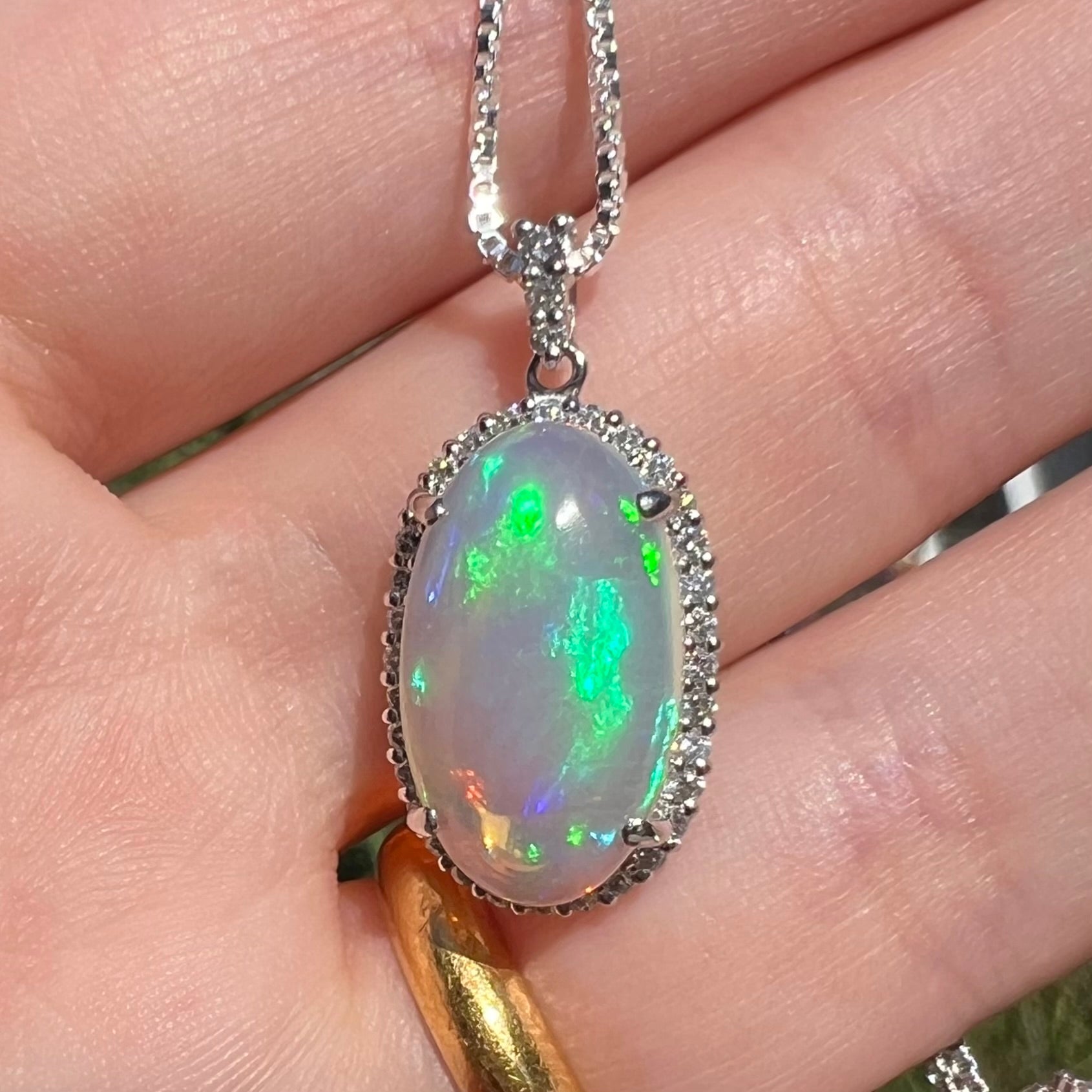 Close-up of a jelly opal and diamond pendant made from platinum, held in a hand in sunlight.  The opal has rolling green and blue play of color.