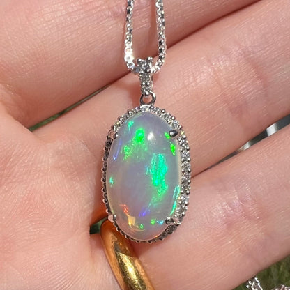 Close-up of a jelly opal and diamond pendant made from platinum, held in a hand in sunlight.  The opal has rolling green and blue play of color.