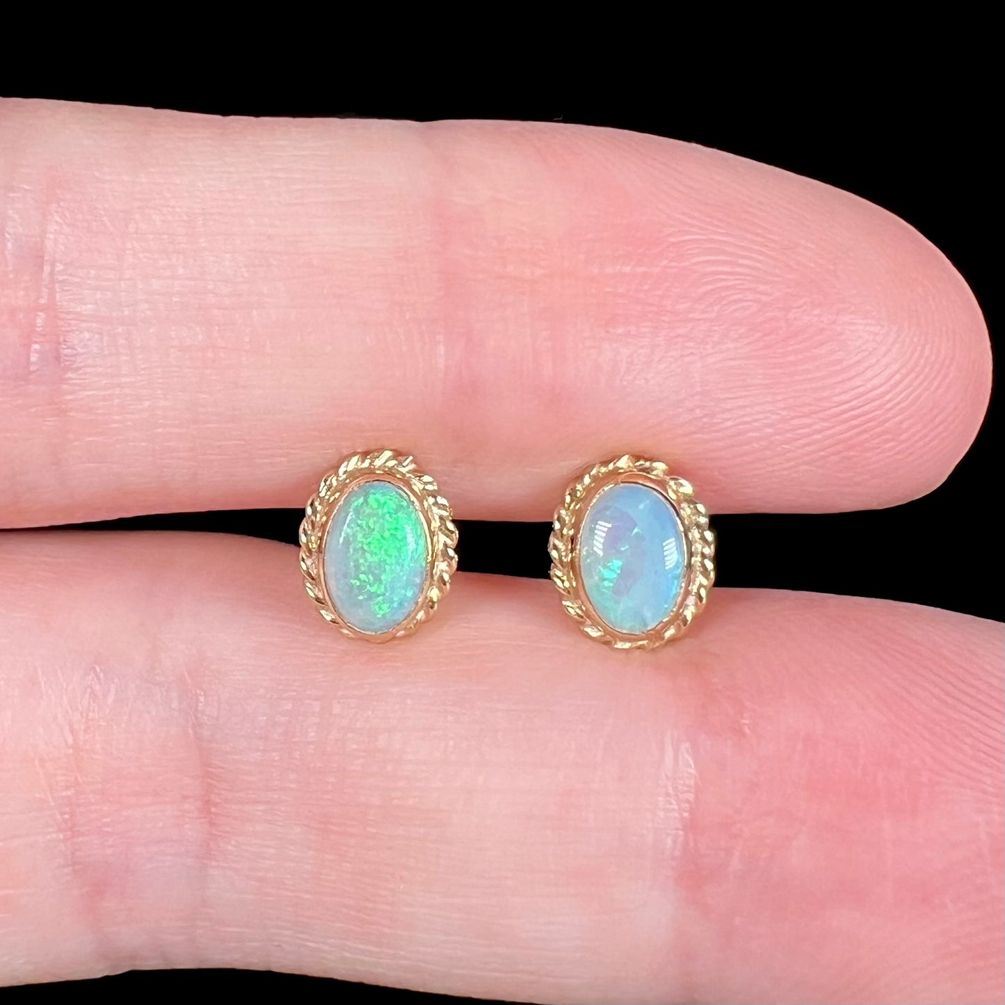 Close-up of green Australian opal stud earrings held between two fingers against black background under LED studio light.