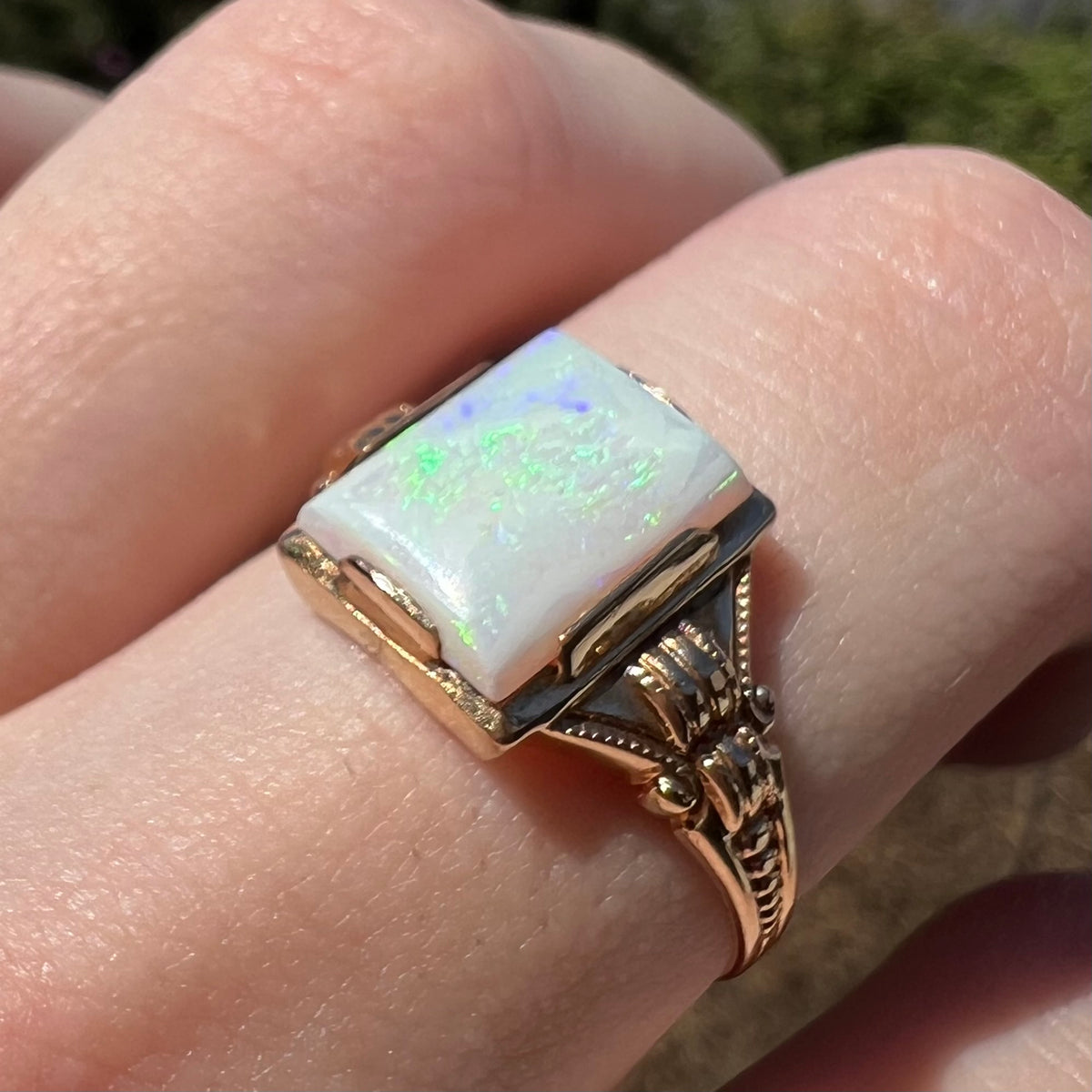 Green and blue flashes seen in a rectangle cut Australian opal, mounted in a 10k gold Art Deco ring setting, worn on a finger in sunlight