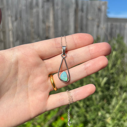 Hand holding a sterling silver opal solitaire necklace in sunlight against outdoor background.  A corner of the opal has soft green play of color.