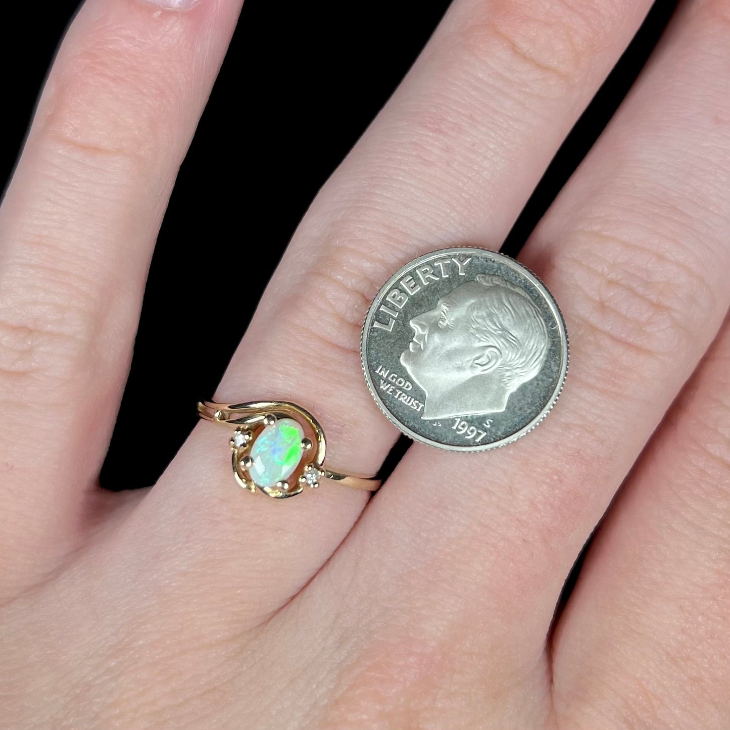 A green, oval-cut Australian opal and diamond ring in yellow gold, worn on a hand next to a US dime coin for scale.