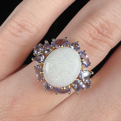Close-up of an oval shaped Australian white opal ring with cluster of tanzanite accents worn on a finger; the opal has green pinfire pattern