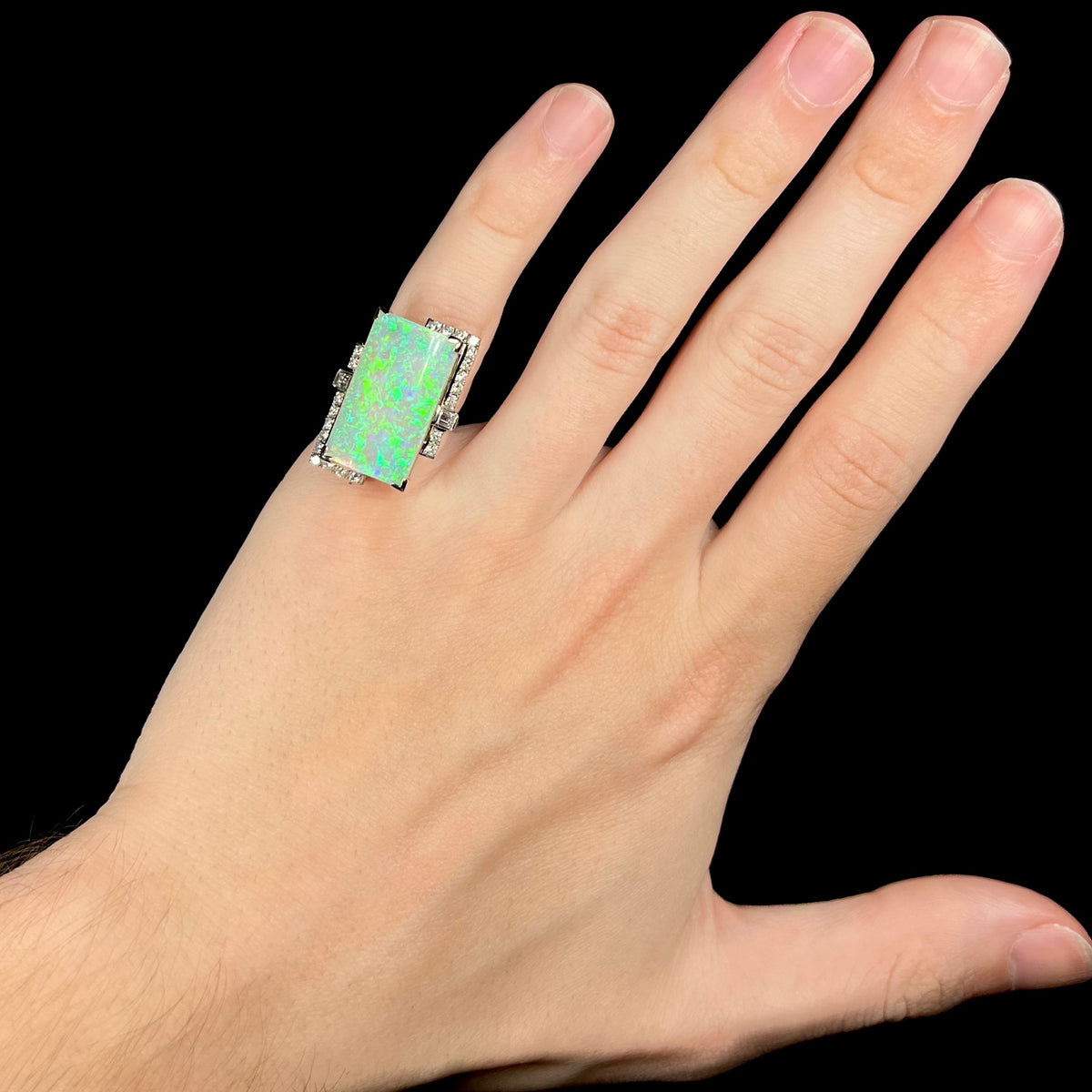 Hand wearing a large, rectangular shaped Australian opal cocktail pinky ring framed by diamonds, showing green play-of-color in opal under cool studio LED light.