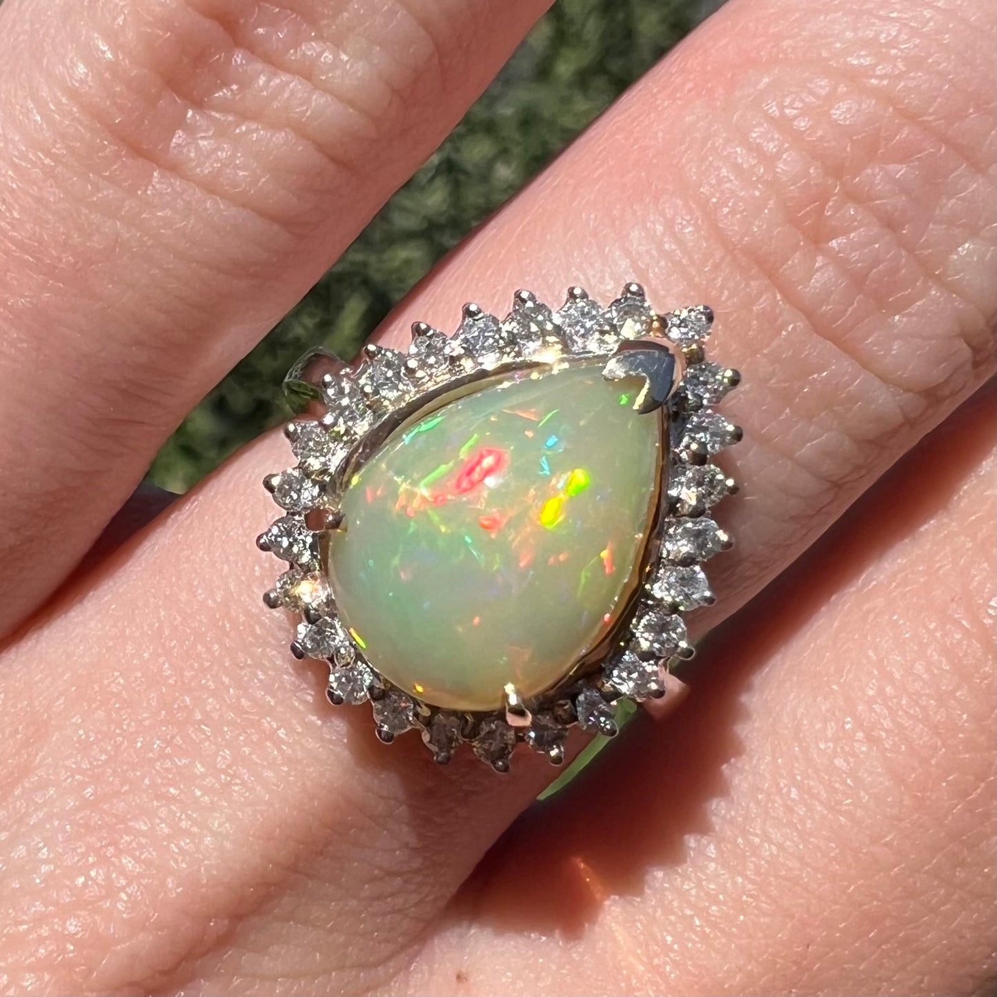 Close-up of a pear shaped Ethiopian opal and diamond engagement ring worn on  a finger in sunlight.  The opal has a greenish body tone.